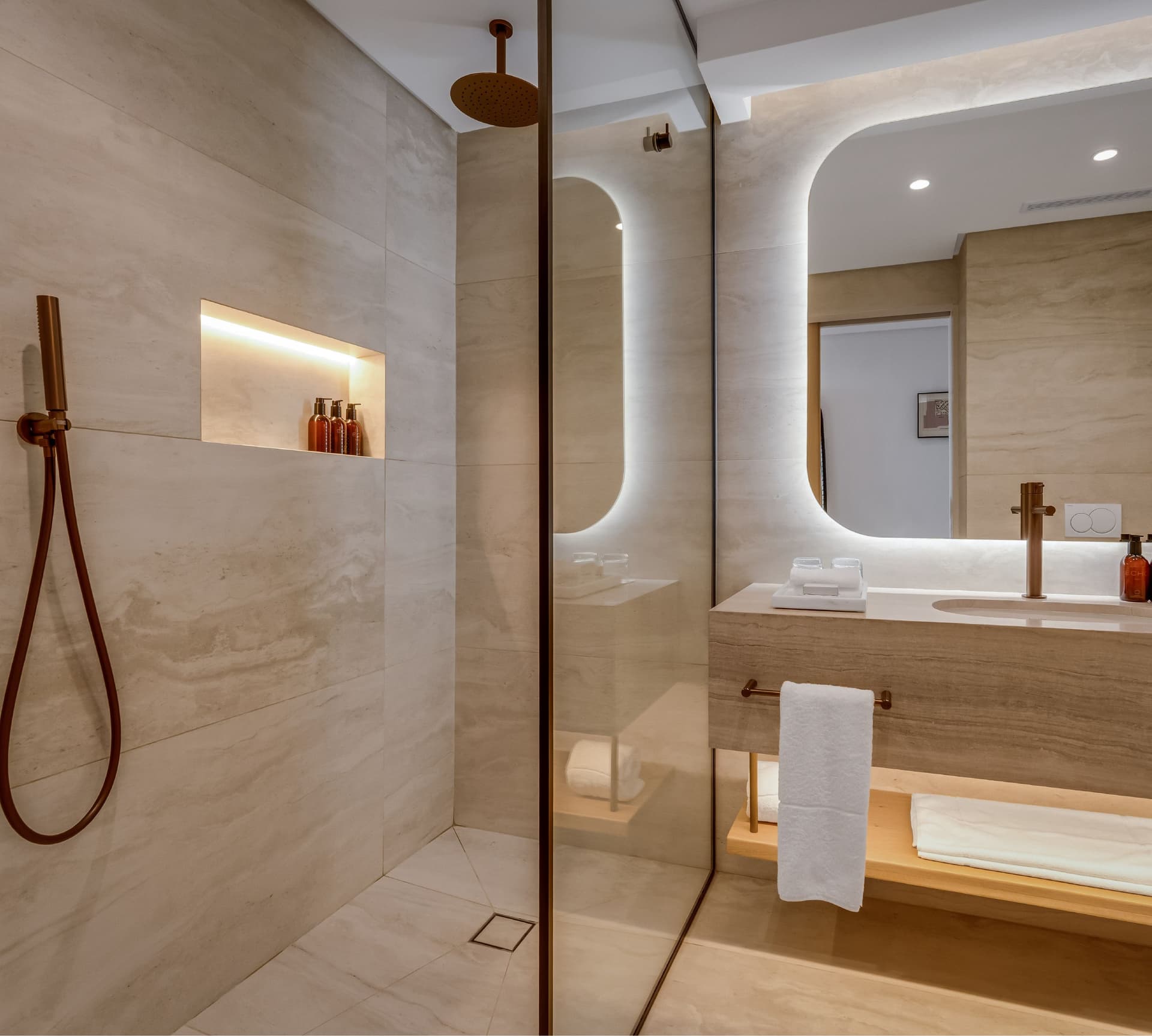 Superior Mountain-Facing Room bathroom at The Cole featuring a glass-enclosed rain shower with rose gold overhead and handheld fittings, illuminated niche with amenities, backlit oval mirror, stone vanity, and timber shelf with white towels