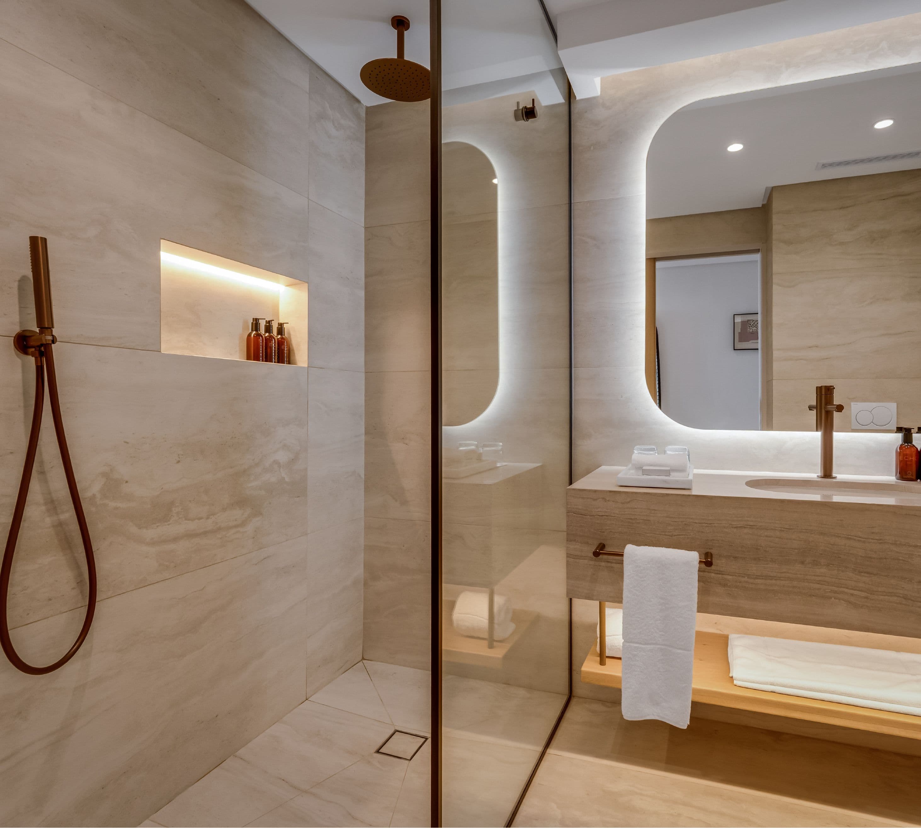 Superior Mountain-Facing Room bathroom at The Cole featuring a glass-enclosed rain shower with rose gold overhead and handheld fittings, illuminated niche with amenities, backlit oval mirror, stone vanity, and timber shelf with white towels