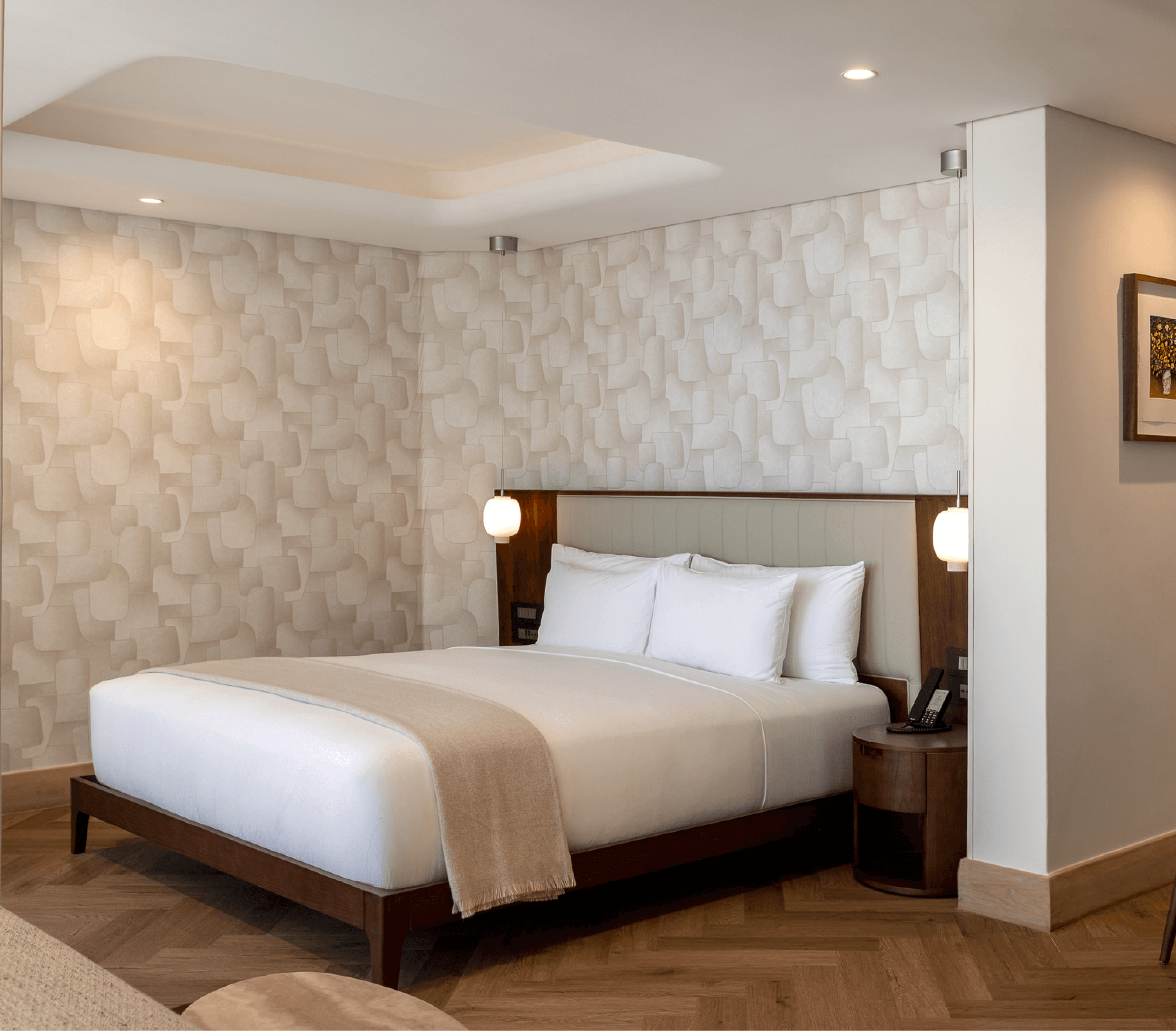 Hotel room with a king bed against a textured cream headboard, flanked by pendant lights, set on herringbone timber flooring with patterned wallpaper behind.
