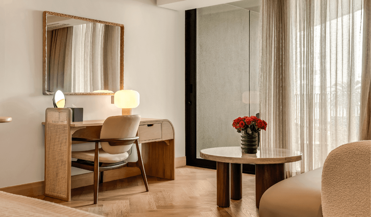 Classic Sea-Facing Room at The Cole featuring a cane-panelled wardrobe, wooden writing desk with table lamp, bouclé armchair, and sheer curtained balcony doors with natural light