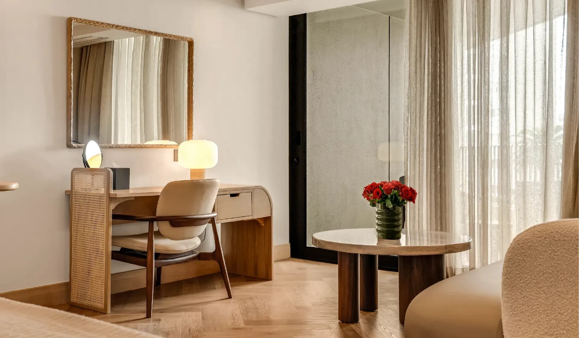 Classic Sea-Facing Room at The Cole featuring a cane-panelled wardrobe, wooden writing desk with table lamp, bouclé armchair, and sheer curtained balcony doors with natural light