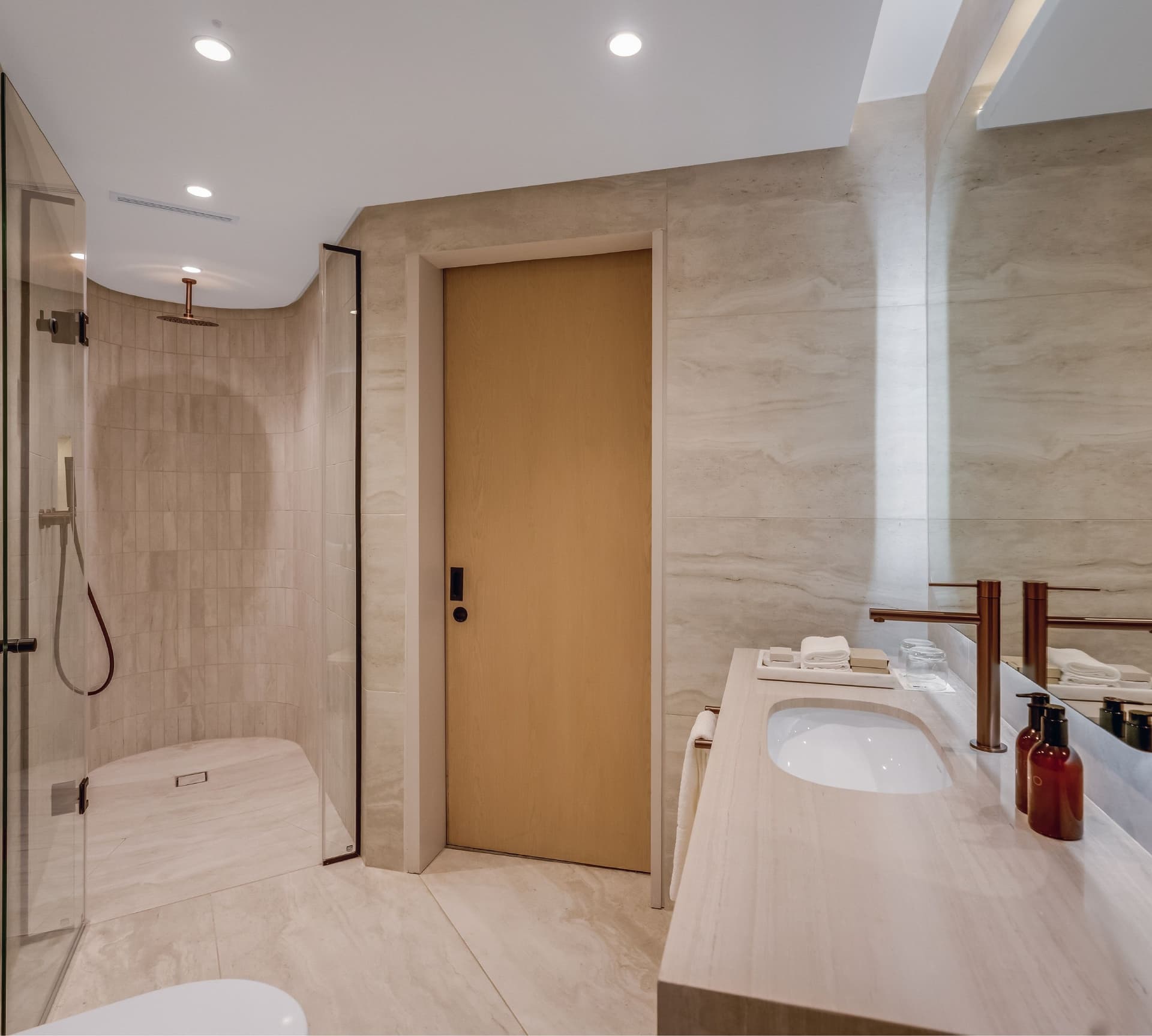 Classic Sea-Facing Room bathroom at The Cole showing a glass-enclosed rain shower, stone basin vanity with rose gold tap, Kove Collection Echo amenities, and warm travertine wall tiles