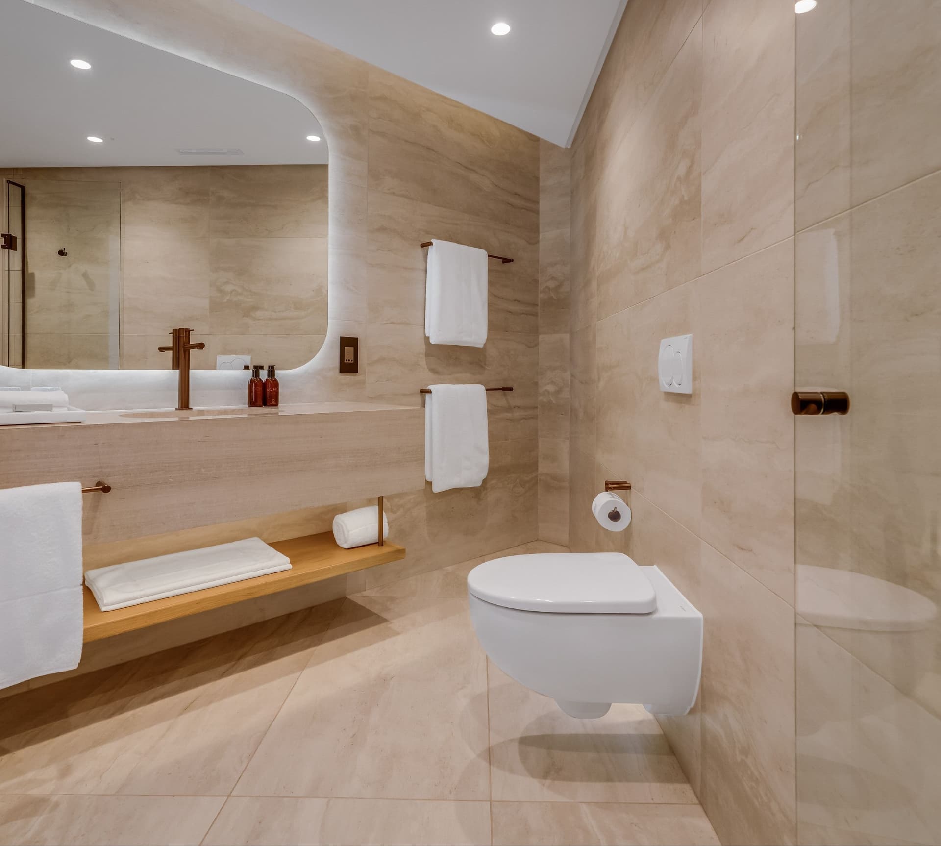 Classic Sea-Facing Room ensuite bathroom at The Cole with travertine-look tiling, backlit rounded mirror, rose gold fixtures, white floating toilet, and neatly folded white towels on a timber shelf