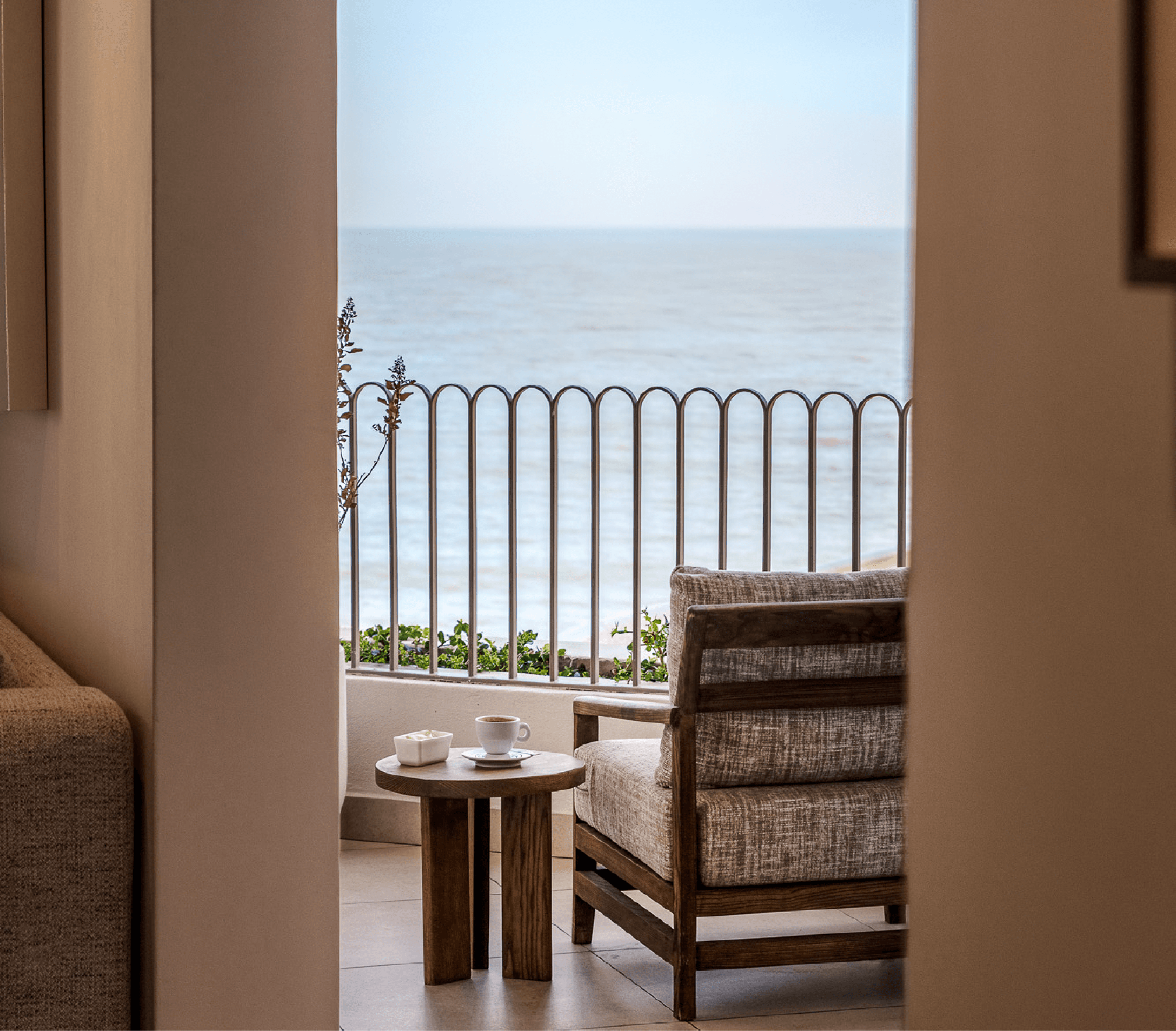 View through an open doorway onto a private balcony with a wood-framed armchair, a round side table set with coffee and butter, a scalloped metal railing, and the open ocean beyond.
