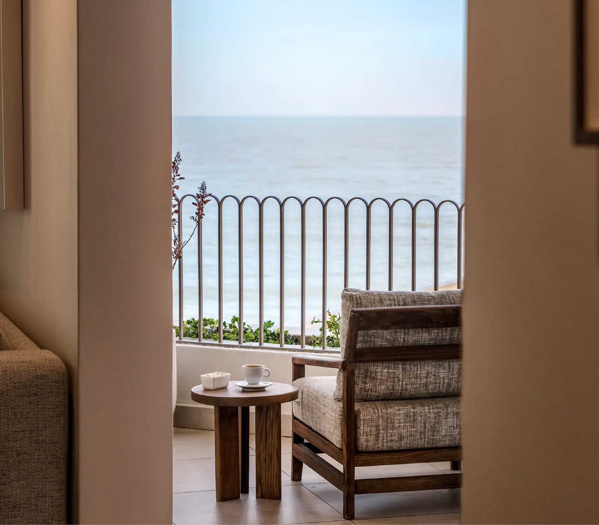 View through an open doorway onto a private balcony with a wood-framed armchair, a round side table set with coffee and butter, a scalloped metal railing, and the open ocean beyond.