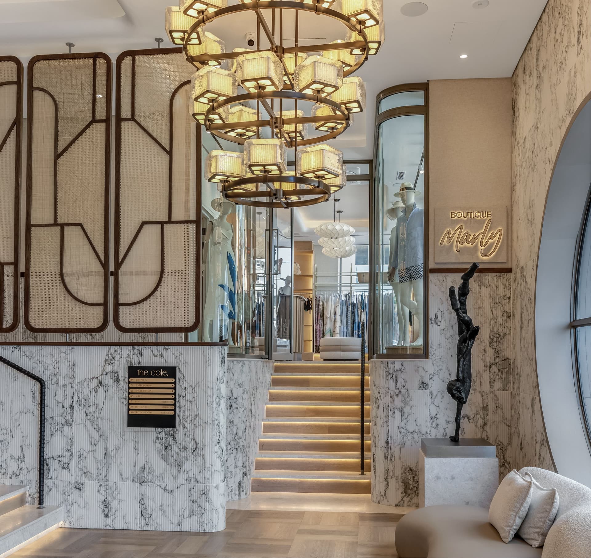 Boutique Marly entrance with a tiered brass chandelier, marble staircase and clothing displays visible beyond the glass storefront
