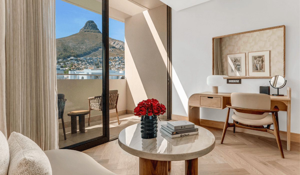 Superior Mountain-Facing Room at The Cole featuring a marble coffee table with red roses and art books, wooden writing desk, and open balcony doors with a direct view of Lion's Head, Cape Town