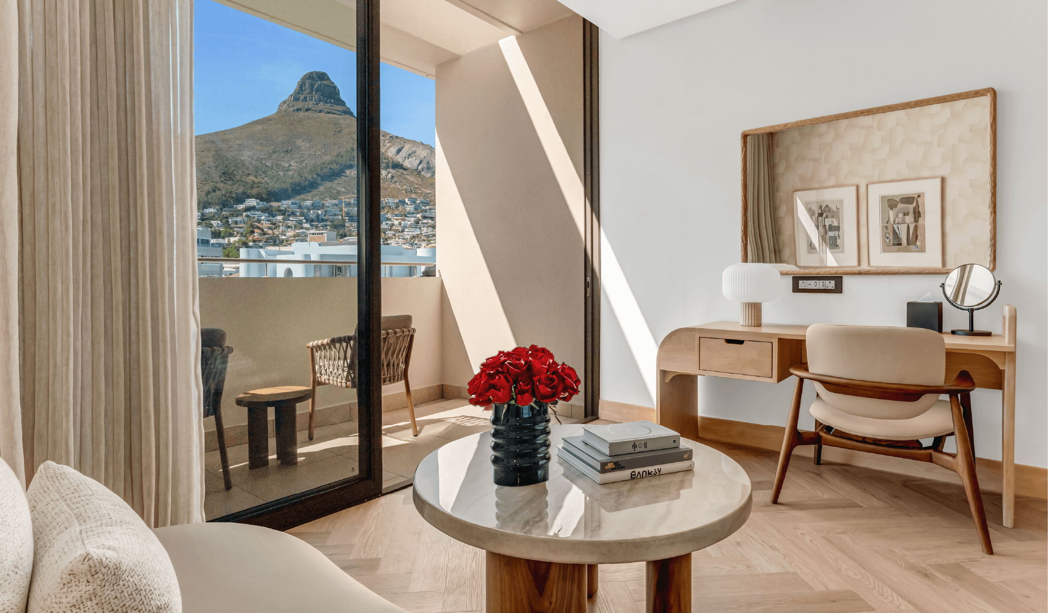 Superior Mountain-Facing Room at The Cole featuring a marble coffee table with red roses and art books, wooden writing desk, and open balcony doors with a direct view of Lion's Head, Cape Town