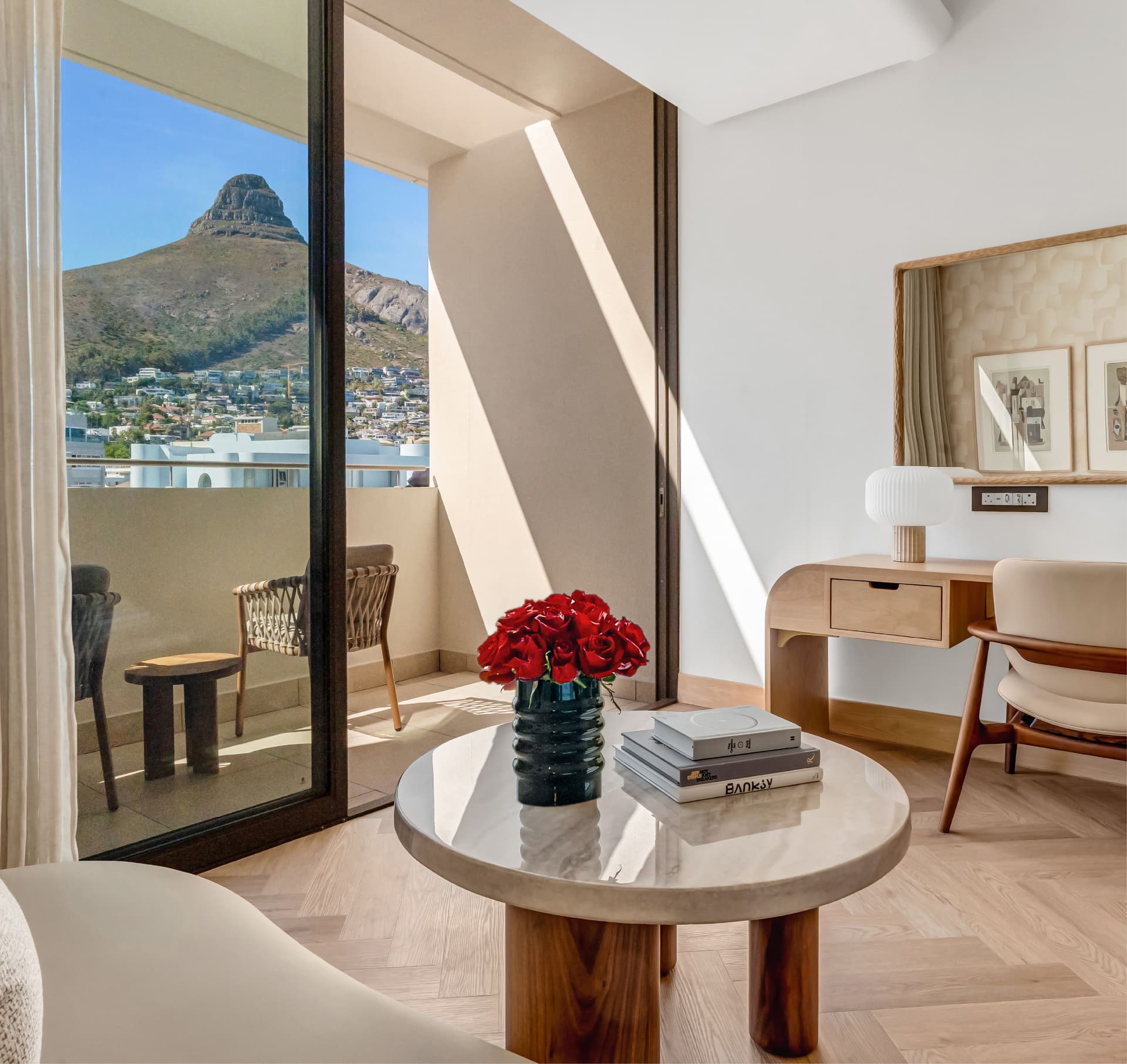 Superior Mountain-Facing Room at The Cole featuring a marble coffee table with red roses and art books, wooden writing desk, and open balcony doors with a direct view of Lion's Head, Cape Town