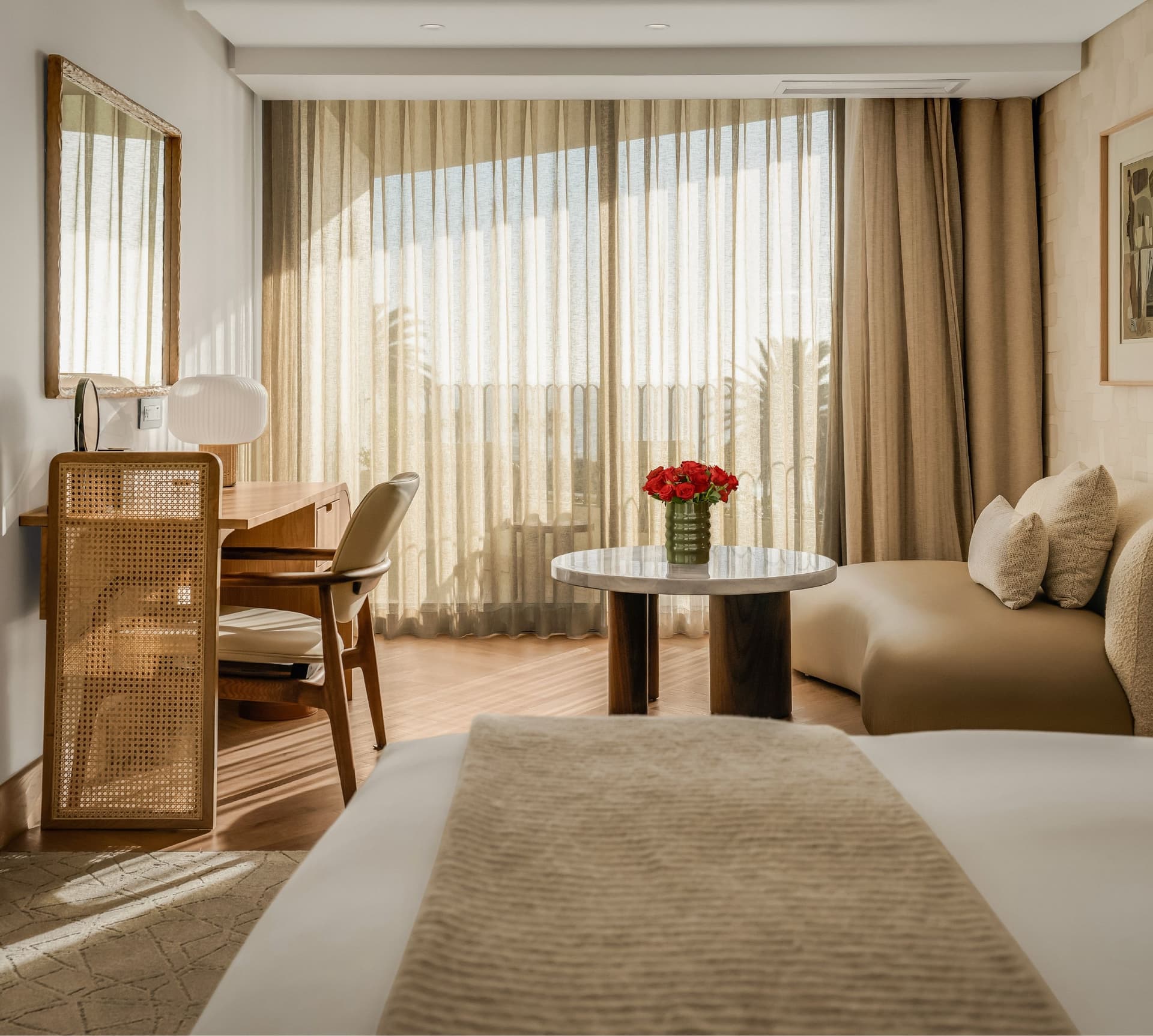 Superior Sea-Facing Room at The Cole with king bed, cane-panelled wardrobe, wooden writing desk, marble coffee table with red roses, curved bouclé sofa, and floor-to-ceiling sheer curtains with palm tree and sea views