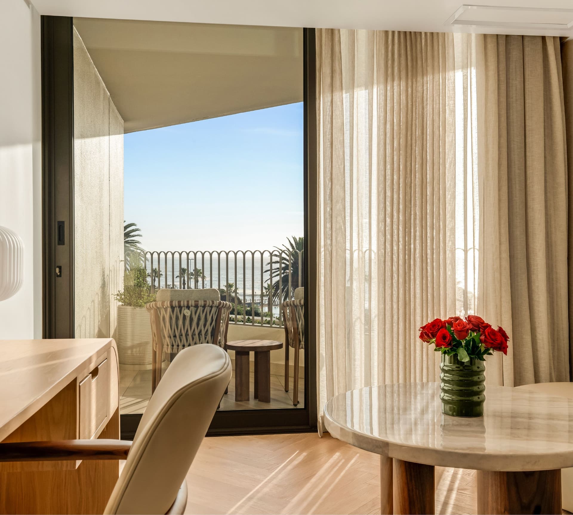 Superior Sea-Facing Room at The Cole featuring a wooden writing desk, marble side table with red rose arrangement, sheer linen curtains, and open balcony doors overlooking palm trees and the sea