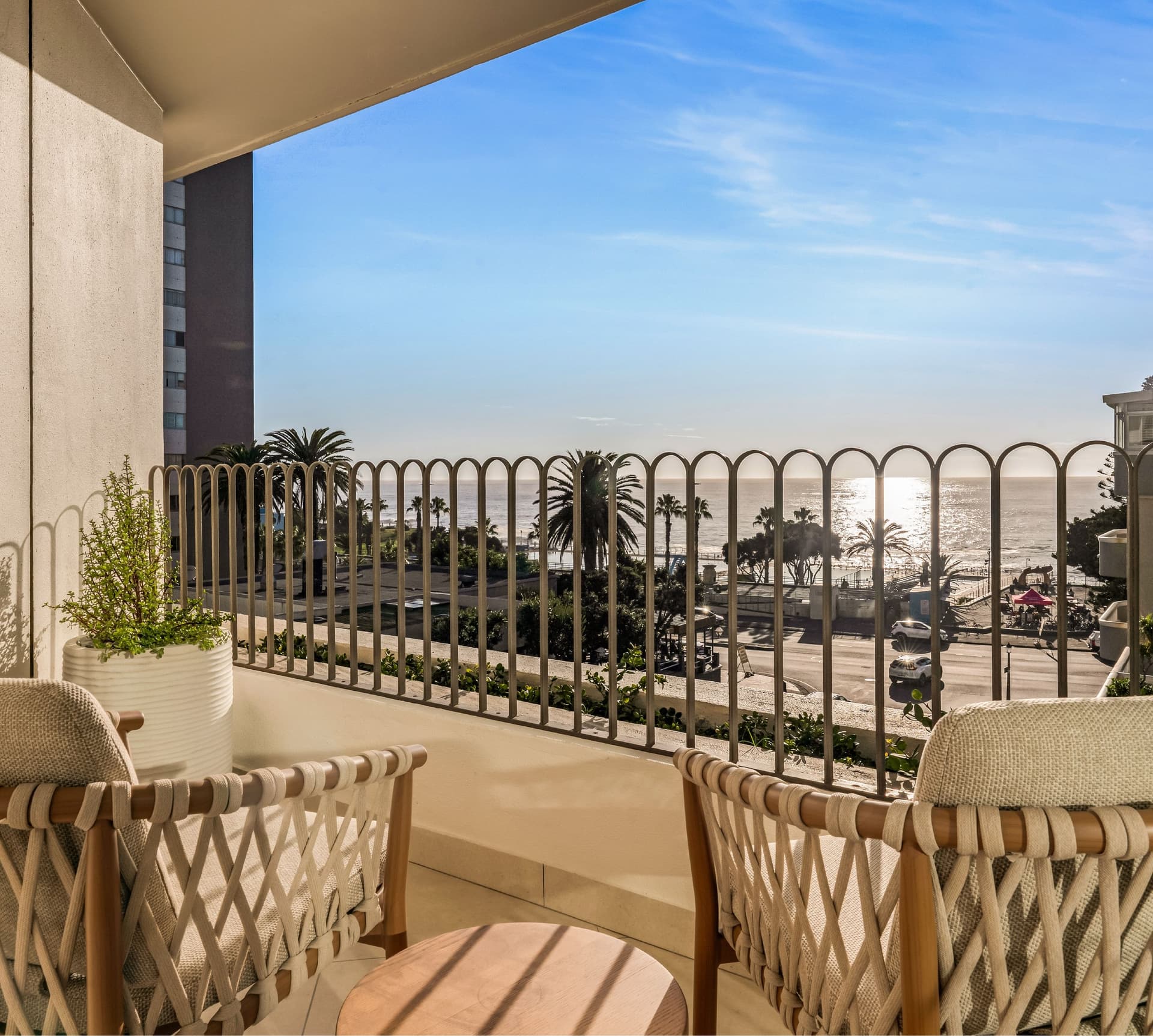Superior Sea-Facing Room private balcony at The Cole with woven rope chairs, potted plant, and panoramic views over Sea Point promenade, palm trees, and the Atlantic Ocean on a clear sunny day